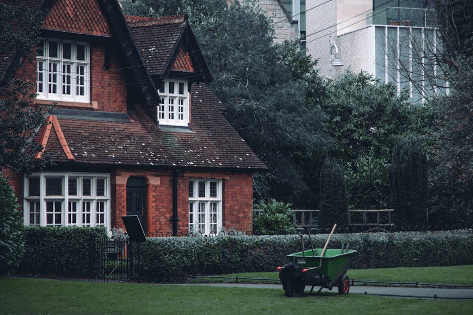 A picturesque garden scene in front of a traditional red-brick house with a steep, tiled roof and white-framed casement windows. The garden features neatly trimmed hedges bordering a well-maintained l