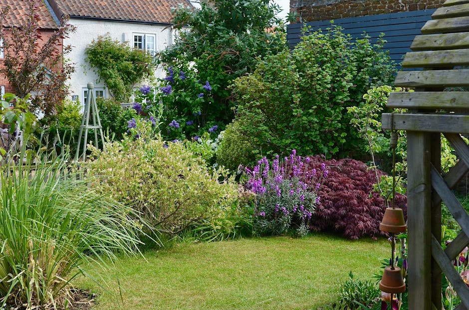 A small front garden with a neatly maintained grassy lawn bordered by various flowering plants and shrubs. On the left, tall green plants with long, narrow leaves are visible. Towards the center, ther